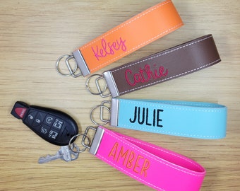 May include: Four personalized leather keychains with metal rings. The keychains are in different colors: orange, brown, light blue, and pink. The names "Kelsey", "Cathie", "Julie", and "Amber" are embroidered on the keychains in pink.