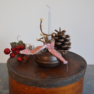 May include: A brass candle holder with a white candle and a red and white ribbon. The candle holder is sitting on a wooden box with a brown lid. There are red berries and a pine cone in the background.