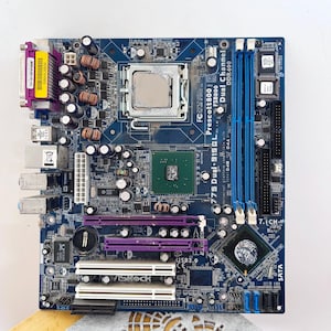 Puede incluir: Una placa base azul ASRock 775 Dual-915GL que presenta un zócalo de procesador central, dos ranuras de memoria DDR400 azules, dos ranuras PCI Express moradas, dos ranuras PCI blancas y varios condensadores, disipadores y puertos integrados.