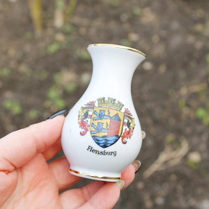 Puede incluir: Jarrón de cerámica blanca con borde dorado y un escudo de armas colorido que presenta un castillo y tres leones. El jarrón tiene la palabra "Flensburg" impresa en la parte delantera.