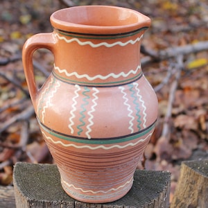 Puede incluir: Una jarra de cerámica marrón con diseños pintados en blanco y verde. La jarra tiene una boca ancha y un asa.