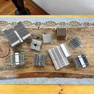Op de afbeelding: Een verzameling van diverse zilver- en koperkleurige computerkoelplaten, met vin-ontwerpen. De componenten zijn gerangschikt op een houten oppervlak, met verschillende vormen en maten voor elektronische koeling.