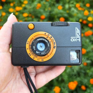 Könnte beinhalten: Eine schwarze Agat 18 Filmkamera mit einem orangefarbenen, kreisförmigen Objektivring mit Blenden- und Verschlusszeitenmarkierungen, einem orangefarbenen Auslöser und einer schwarzen Handschlaufe. Das Objektiv ist mit Industar-104 und 2,8 beschriftet.