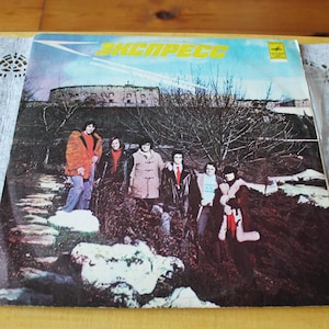 May include: Vintage vinyl record album cover featuring a band of six men in coats and jackets standing in front of a building. The album title is in yellow text against a blue sky background.