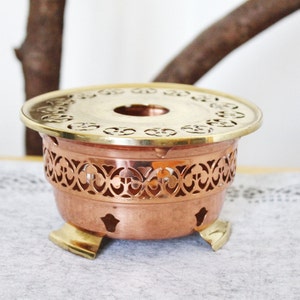 Può includere: Un scaldavivande per tè in rame e ottone con un design decorativo tagliato. Lo scaldavivande ha tre gambe e una parte superiore rotonda con un foro al centro.