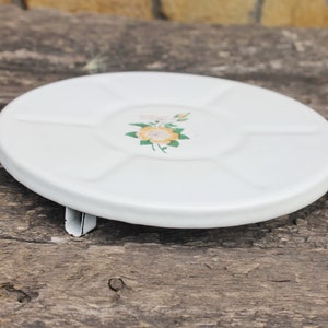 Può includere: Un sottopentola in ceramica bianca con un design floreale che presenta un fiore giallo con foglie verdi. Il sottopentola ha tre gambe.