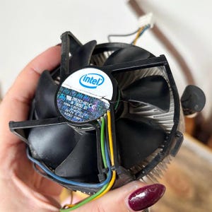 May include: A black Intel CPU fan with a silver heat sink. The fan has four curved blades and a central Intel logo. The fan has a sticker with the text "096263-001 CE" and other technical details. Wires in blue, green, and yellow extend from the fan.