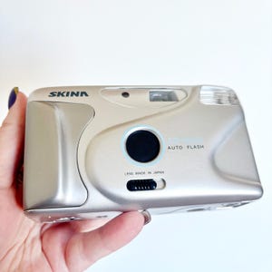 Puede incluir: Cámara SKINN plateada con lente negra y botón negro. La cámara tiene las palabras "AUTO FLASH" y "LENS MADE IN JAPAN" impresas. La cámara está sujeta por una persona.