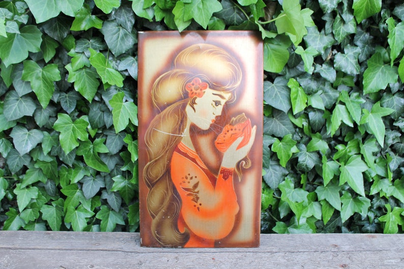 May include: A vintage wall art piece featuring a woman with long brown hair holding a red flower. The woman is wearing a long orange dress with gold accents. The background is a gold and brown color.