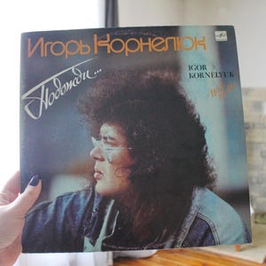 May include: A vintage vinyl record album cover featuring a portrait of Igor Kornelyuk, a musician with a bushy afro, wearing glasses and a denim jacket. The album title is "Новогодняя" in Cyrillic script.