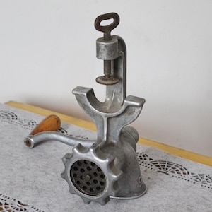May include: Vintage silver-toned meat grinder with a wooden handle. The grinder has a clamp for securing it to a surface and a circular grinding plate. The handle is made of wood. The grinder is designed for manual operation.