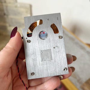 Pode incluir: Um pequeno componente de alum&iacute;nio escovado com uma ventoinha e um pequeno autocolante que diz "Brushless". O componente tem uma forma quadrada com cantos arredondados e &eacute; segurado por uma m&atilde;o com verniz de unhas roxo escuro.