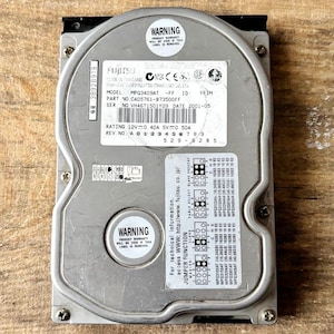 Puede incluir: Un disco duro Fujitsu plateado con múltiples etiquetas y texto, incluyendo "ADVERTENCIA" y especificaciones técnicas. El disco es rectangular con bordes redondeados y agujeros para tornillos. El texto incluye números de modelo y serie.