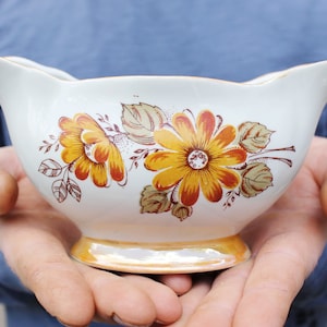 May include: A white ceramic gravy boat with a floral design featuring yellow, orange, and brown flowers. The boat has a scalloped edge and a smooth, glossy finish.
