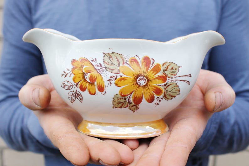 May include: A white ceramic gravy boat with a floral design featuring yellow, orange, and brown flowers. The boat has a scalloped edge and a smooth, glossy finish.