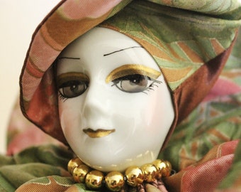 May include: A porcelain doll with a painted face and a colorful fabric hat and costume. The doll has brown eyes, a pink mouth, and a gold necklace.