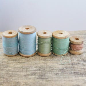 May include: Five vintage wooden spools of thread in varying sizes and colors. The thread colors include light blue, green, and peach. The spools are arranged on a weathered wooden surface, showcasing a rustic aesthetic.