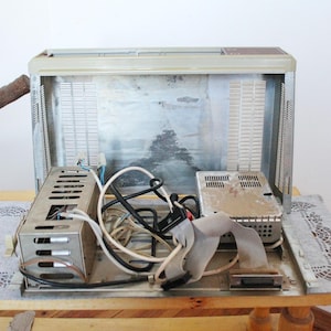 May include: An open, silver-coloured metal box with wires and electronic components inside. The box has a metal grate on the front and a metal panel on the back. The box is sitting on a wooden surface.