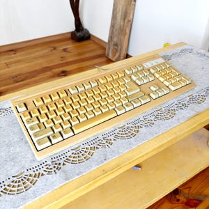 May include: A vintage beige computer keyboard with off-white keys. The keyboard has a rectangular shape and a wired connection. It is placed on a light gray, lacy patterned mat on a wooden surface.