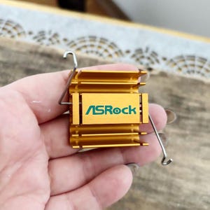 May include: A gold-colored computer component with the text "ASRock" in green. The component has a rectangular shape with multiple fins and is held by a silver wire clip. The background is blurred.