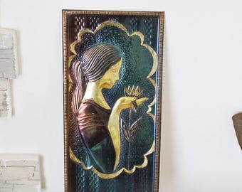 May include: A vintage wall sculpture featuring a woman in relief, with a gold finish, holding a flower. The sculpture is set against a dark green and blue background with a metallic sheen.