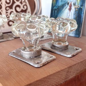May include: A set of clear glass door knobs with a rounded, textured surface and a scalloped edge. Each knob is attached to a rectangular, weathered silver-toned metal base plate featuring four small screw holes for mounting purposes.