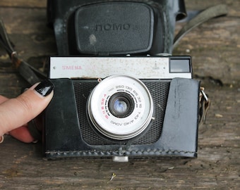 May include: A black and silver vintage film camera with the text "SMENA" on the front. The camera has a black leather case and a silver lens with a black ring around it. The lens has numbers printed on it, including "1:2.8/40" and "250 130 85 32 16 8 4".