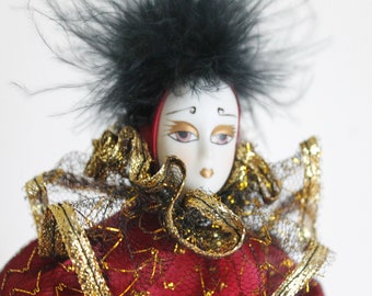 May include: A porcelain doll with a black feather headdress, red and gold costume, and a painted white face with black eyes and red lips.