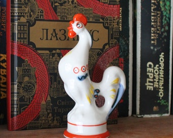 May include: A white ceramic rooster figurine with a red comb and beak, blue and yellow accents, and a red and orange base. The figurine is displayed on a wooden shelf in front of a row of books.
