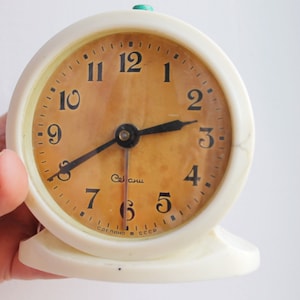 Pode incluir: Relógio despertador vintage branco com ponteiros e números pretos. O mostrador do relógio é de cor castanho claro com a palavra "СССР" impressa na parte inferior.