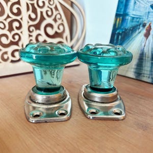 May include: Two matching door knobs featuring translucent teal glass tops with a circular, scalloped edge design. Each knob is mounted on a rectangular, brushed silver-toned metal base plate with two pre-drilled screw holes for installation.