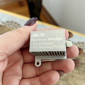 Puede incluir: Componente plateado ASUS sin ventilador con el texto "Fanless Design - Zero Noise - No Moving Parts". El componente tiene una serie de aletas paralelas y una pequeña pieza de plástico blanco.