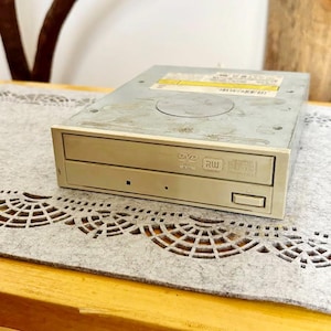 May include: A beige DVD-RW drive with a rectangular shape. The drive has a slot for inserting discs and a button for ejecting them. The front of the drive has the DVD and RW logos. The drive is sitting on a gray, patterned surface.