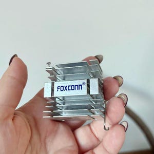 May include: A silver-colored heat sink with the word "FOXCONN" printed in blue. The heat sink has multiple parallel fins and is held in a hand.