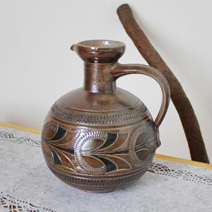 Könnte beinhalten: Eine braune Keramikkanne mit einer breiten Öffnung und einem Henkel. Die Kanne hat ein dekoratives Design aus schwarzen und braunen Blättern und Wirbeln.