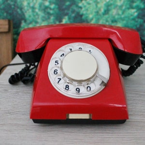 Puede incluir: Un teléfono rojo vintage de disco rotatorio con un dial blanco y detalles en negro. El teléfono tiene un diseño clásico con un auricular y un cable en espiral. El teléfono está sobre una superficie de color claro.