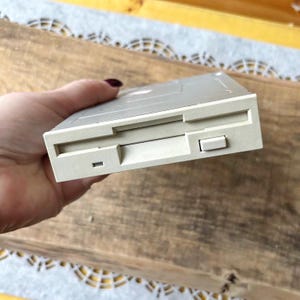 Puede incluir: Una unidad de disquete beige de 3,5 pulgadas, un dispositivo de almacenamiento informático antiguo. La unidad tiene forma rectangular con una ranura para la inserción del disco y un botón. El dispositivo se sostiene en una mano.