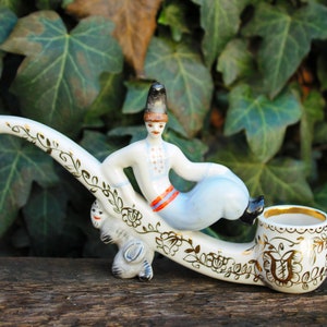 May include: A white porcelain pipe with a gold floral design. A figurine of a man in traditional clothing sits on the pipe, holding a pipe.