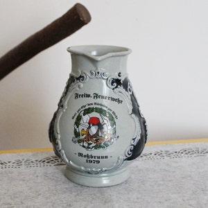 Puede incluir: Una jarra de cerámica blanca con detalles azules y dorados. Presenta una representación de un bombero con un casco rojo y el texto "Freiw. Feuerwehr" y "Gott zur Ehr, dem Nächsten zur Wehr -  Rohrbunn - 1979".
