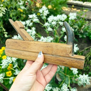 Pode incluir: Uma plaina manual de madeira vintage com corpo retangular de acabamento em veios naturais, uma cunha de metal para a lâmina inclinada no topo e um cabo escuro curvo fixado na extremidade traseira para trabalhos manuais em madeira.