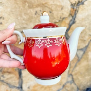 Puede incluir: Una tetera de porcelana con un cuerpo rojo vibrante y una tapa, asa y boquilla blancas. La parte superior del cuerpo rojo está decorada con un patrón floral dorado. La tapa tiene un pequeño pomo dorado y la base es de color blanco.