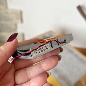 Pode incluir: Um componente de computador prateado com dissipador de calor e ventoinha. O componente tem uma etiqueta com o texto "251-30920-0700F Z1038S-0". Fios vermelhos e pretos est&atilde;o conectados.