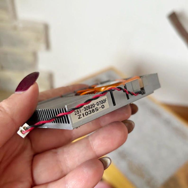 Pode incluir: Um componente de computador prateado com dissipador de calor e ventoinha. O componente tem uma etiqueta com o texto "251-30920-0700F Z1038S-0". Fios vermelhos e pretos est&atilde;o conectados.