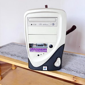 May include: A beige and black computer tower case featuring a CD-ROM drive labeled 52X Max, a purple and white logo reading Impression Computers, a power button, and a small square emblem with a colorful cube graphic on the front panel.