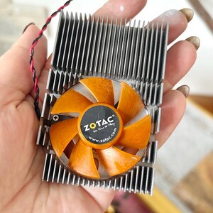 Pode incluir: Um componente de computador com um dissipador de calor prateado e uma ventoinha laranja com o log&oacute;tipo ZOTAC. As p&aacute;s da ventoinha s&atilde;o laranja e o dissipador de calor tem m&uacute;ltiplas aletas verticais. Um fio vermelho e preto est&aacute; ligado.