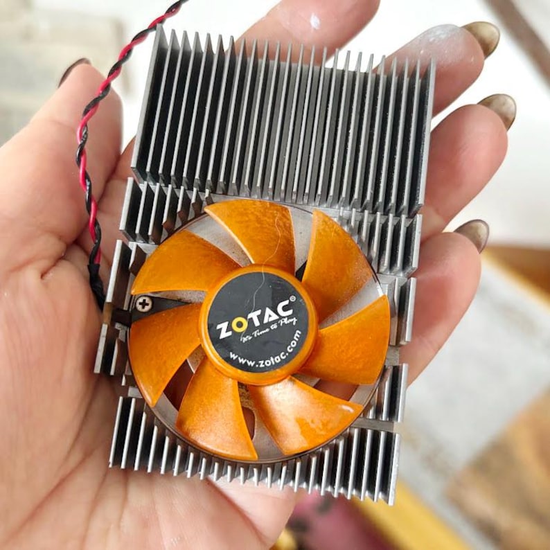 Pode incluir: Um componente de computador com um dissipador de calor prateado e uma ventoinha laranja com o log&oacute;tipo ZOTAC. As p&aacute;s da ventoinha s&atilde;o laranja e o dissipador de calor tem m&uacute;ltiplas aletas verticais. Um fio vermelho e preto est&aacute; ligado.
