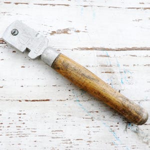 May include: A vintage glass cutter with a wooden handle. The cutter is made of metal and has a silver finish. The handle is brown and has a slightly worn look.