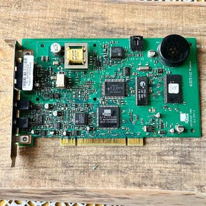 Puede incluir: Una placa de circuito verde con varios componentes electrónicos, incluyendo chips, condensadores y un componente cilíndrico negro. La placa tiene conectores dorados y pequeños componentes negros y plateados. El texto "Com" es visible.