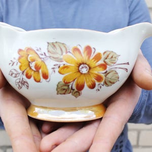 May include: A white ceramic gravy boat with a floral design featuring yellow, orange, and brown flowers. The boat has a gold rim and is being held by a person's hands.