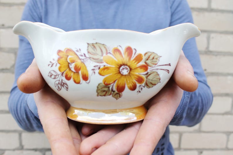 May include: A white ceramic gravy boat with a floral design featuring yellow, orange, and brown flowers. The boat has a gold rim and is being held by a person's hands.